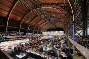 Large indoor car exhibition at an industrial-style venue with many vehicles and visitors.