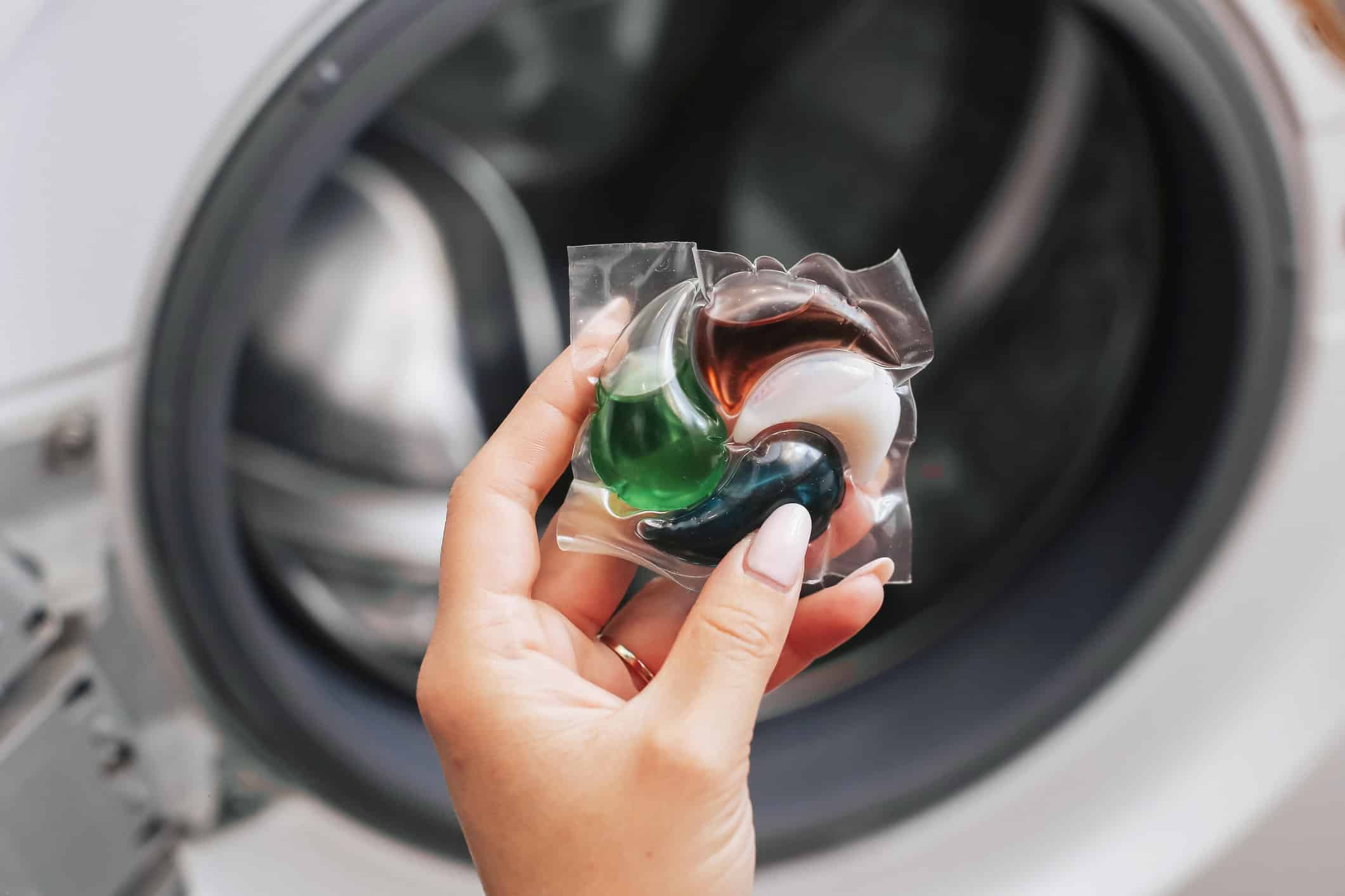 Laundry detergent pod in hand. Capsule for washing machine. Household cleaning product. Liquid laundry detergent pods in various colors held in a hand in front of a washing machine.